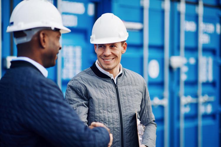 Two men, wearing hardhats, shaking hands in front of shipping containers.