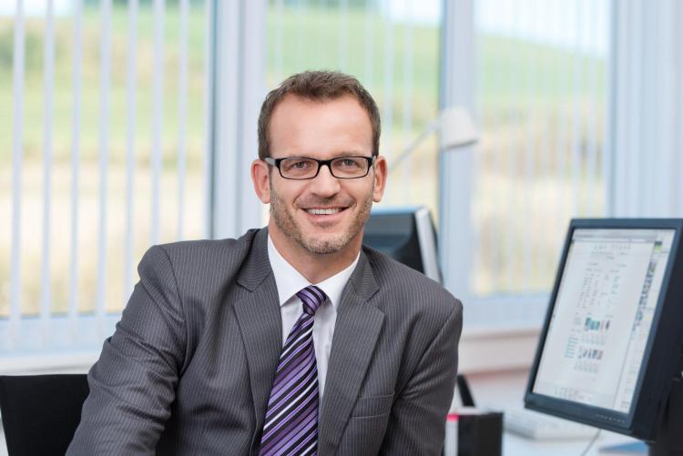 Man in suit sitting at office desk, looking at camera and smiling.