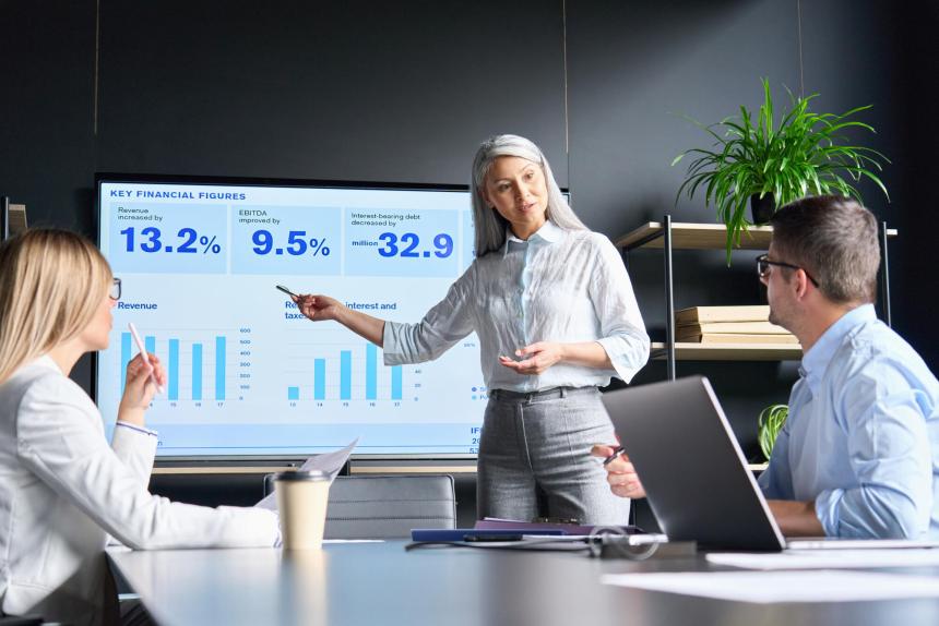 Woman standing in front of digital display leading a meeting. Two people are sitting at a table looking at her.