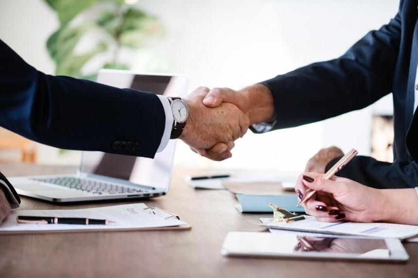 Close up of 2 men shaking hands across a table while a woman takes notes