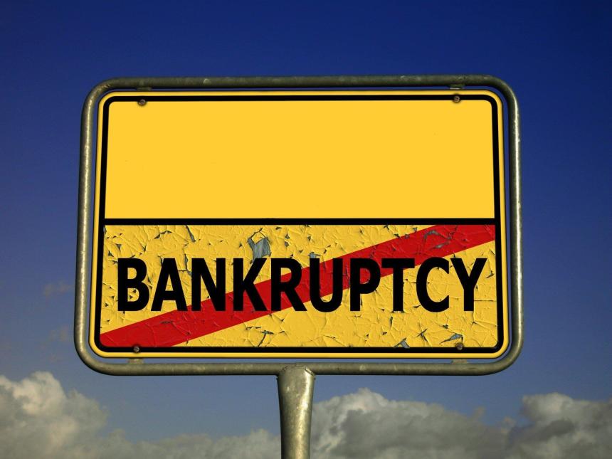 Yellow road sign against blue sky. Sign says bankruptcy in black letters with a red diagonal line over the word.