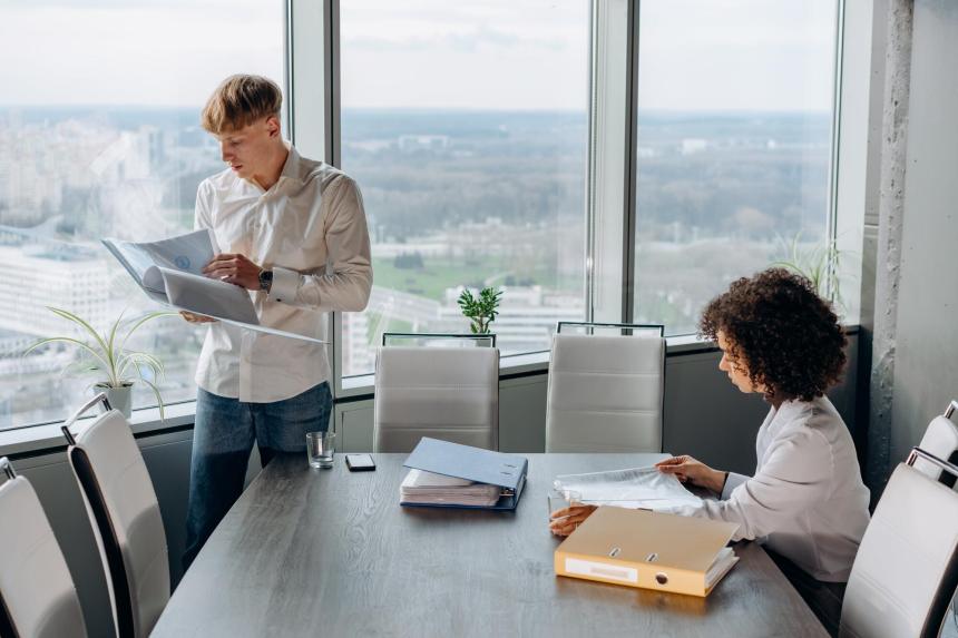 People near an office table, 1 sitting, 1 standing, reviewing paperwork